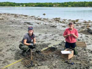 Two students working in the intertidal zone of the Damariscotta River, looking at camera while counting shellfish and crabs.